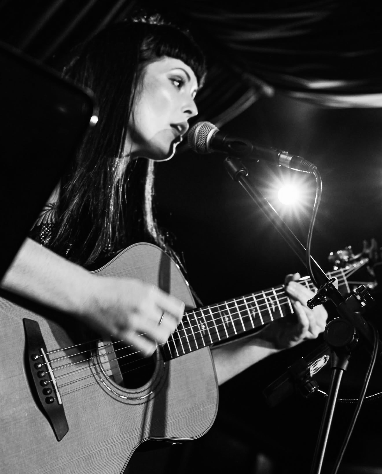 Grace de la Hunt singing into a microphone while playing acoustic guitar on a dimly lit stage